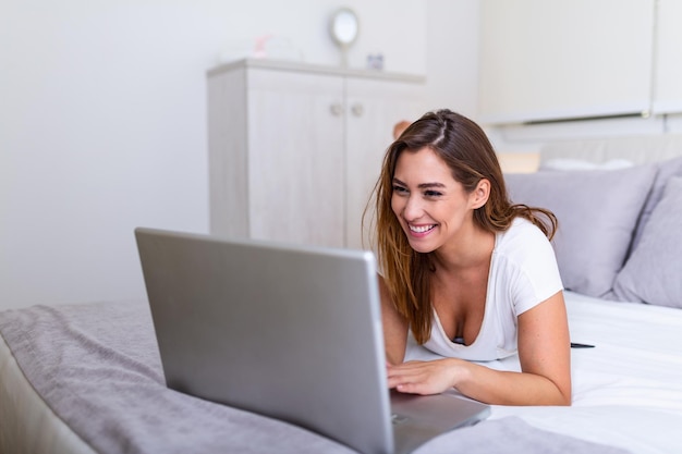 Beautiful young college student doing homework in bed at home, young pretty woman preparing school test in bedroom with laptop pc. Freelance working from home concept