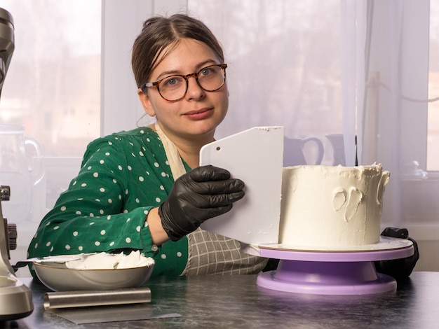 Beautiful young confectioner girl makes a cake with white cream using a cooking spatula