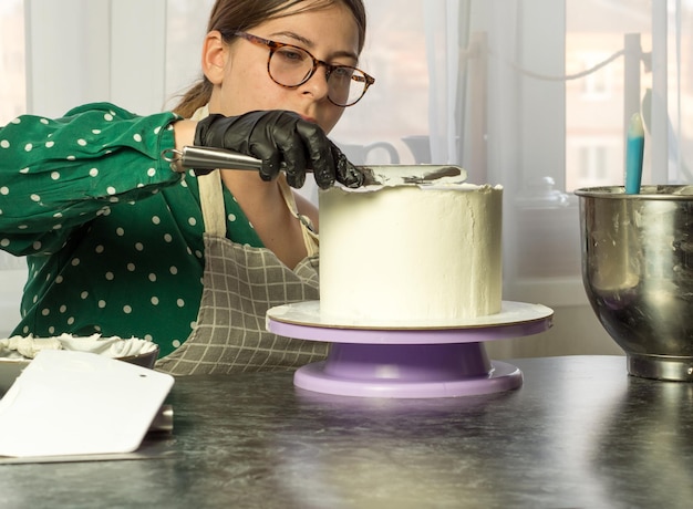 La bella giovane pasticcera fa una torta con crema bianca usando una spatola da cucina