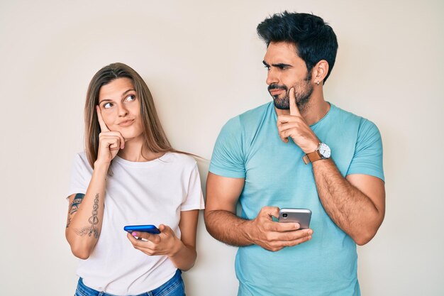 Beautiful young couple of boyfriend and girlfriend using smartphone serious face thinking about question with hand on chin thoughtful about confusing idea