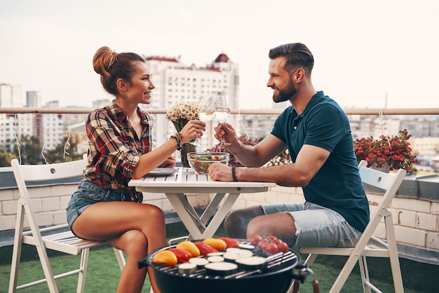 Beautiful young couple in casual clothing enjoying romantic dinner and smiling while enjoying romantic dinner on the rooftop patio outdoors