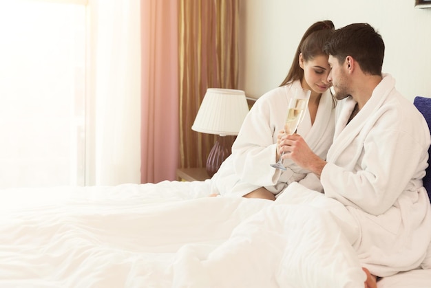 Beautiful young couple enjoying champagne and relaxing in SPA center. Happy man and woman lying in bed with glasses in hotel room, copy space