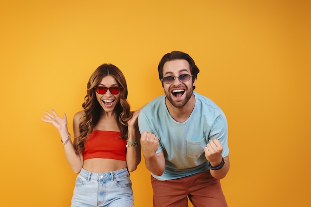 Beautiful young couple in eyeglasses having fun while standing against yellow background