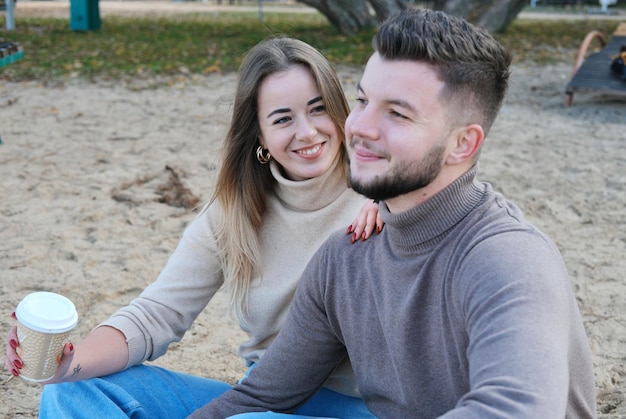 A beautiful young couple a guy and a girl are sitting on the beach and drinking coffee the girl looks at the guy and smiles