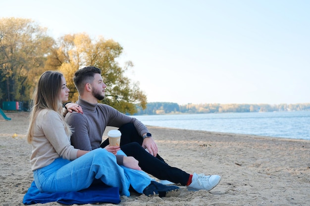 A beautiful young couple a guy and a girl are sitting on the beach and drinking coffee man and woman enjoy lake view