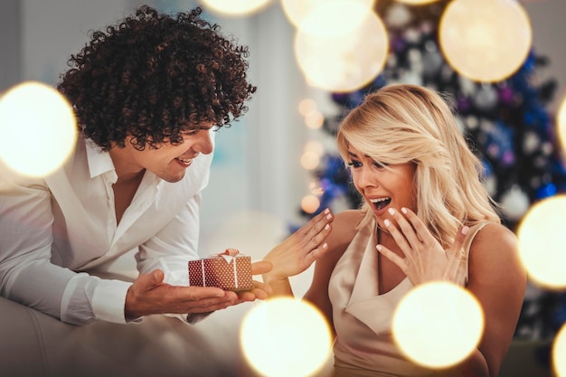 Beautiful young couple is celebrating Christmas at home. Handsome smiling man is giving a gift box to his pretty girlfriend.