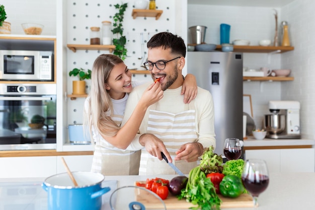 美しい若いカップルはお互いに餌をやり、自宅のキッチンで料理をしながらにこにこ笑っている幸せなカップルは、軽いキッチンで健康的な食べ物を準備しています