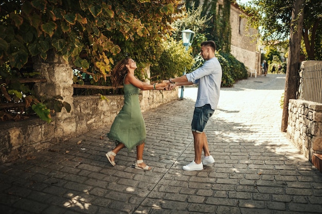 A beautiful young couple is having fun and dancing on the street while walking along of a Mediterranean town. They are enjoyed in summer sunny day, holding each other's hand and smiling.