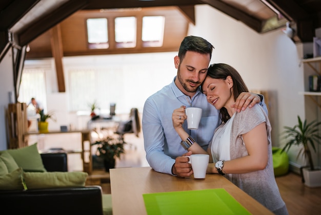 Beautiful young couple is hugging and smiling while drinking tea in kitchen at home. 