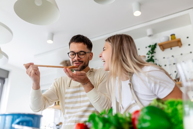 Beautiful young couple is looking at each other and feeding each other with smiles while cooking in kitchen at home Loving joyful young couple embracing and cooking together having fun in the kitchen