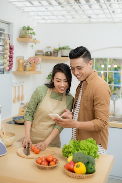 美しい若いカップルは、自宅のキッチンで料理をしながら携帯電話を使用して笑っています