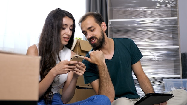 Beautiful young couple just moved in new flat. They are sitting on the floor and searching online for furniture