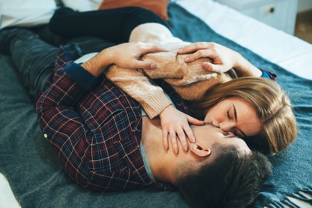 Beautiful young couple kissing and embracing while leaning on the bed at their home .