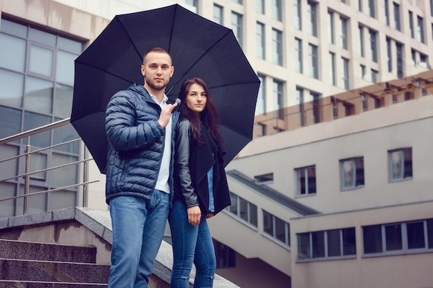 Beautiful young couple in love on a date outdoors on modern urban background Bearded handsome man and brunette pretty woman in casual dress with umbrella Rainy weather