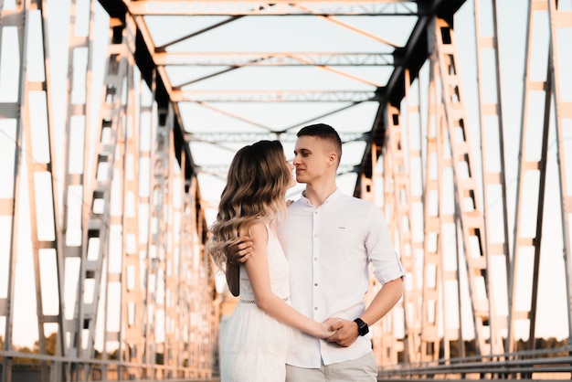 A beautiful young couple in love, a man and a woman, embrace, kiss on a large metal bridge at sunset.