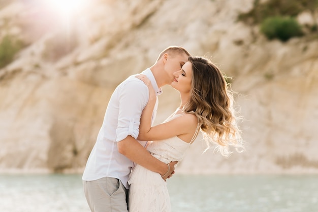 A beautiful young couple in love, a man and a woman embrace, kiss near a blue lake and sand at sunset.