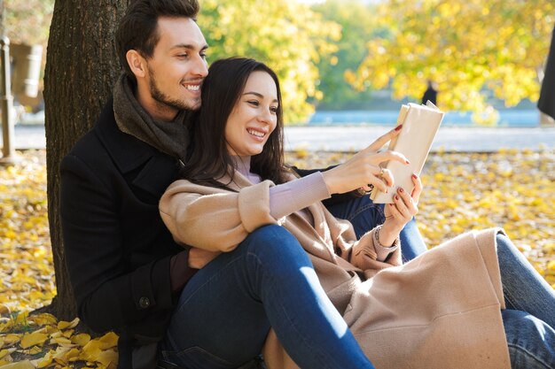 Photo beautiful young couple in love spending time together at the park in autumn, reading a book while sitting under the tree