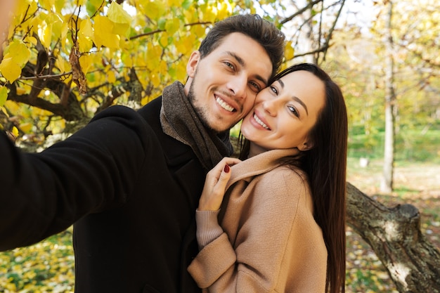 Beautiful young couple in love spending time together at the park in autumn, taking a selfie, hugging