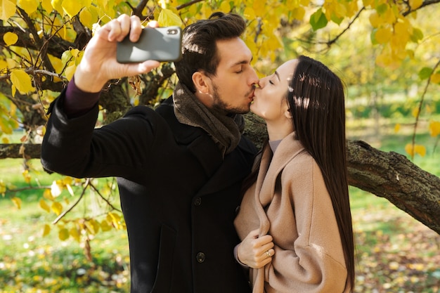 Beautiful young couple in love spending time together at the park in autumn, taking a selfie, kissing