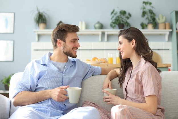 Beautiful young couple in pajamas is looking at each other and smiling on a sofa in the living room.