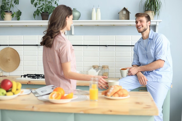 パジャマ姿の美しい若いカップルは、自宅のキッチンで料理をしながらお互いを見て笑っています。