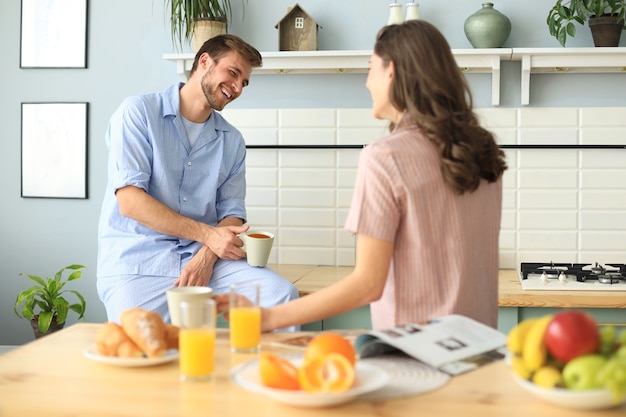 パジャマ姿の美しい若いカップルは、自宅のキッチンで料理をしながらお互いを見て笑っています。