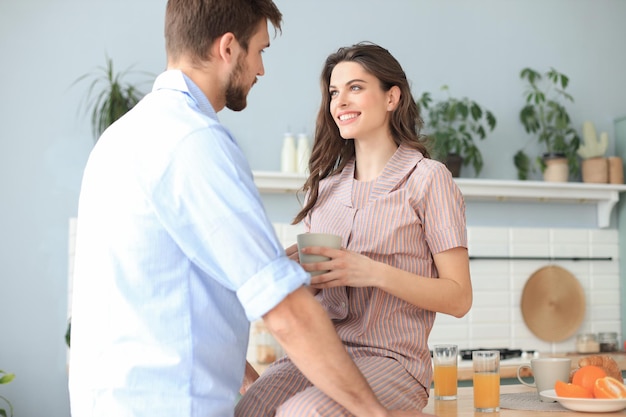 Beautiful young couple in pajamas is looking at each other and smiling while cooking in kitchen at home.