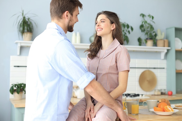 Beautiful young couple in pajamas is looking at each other and smiling while cooking in kitchen at home.