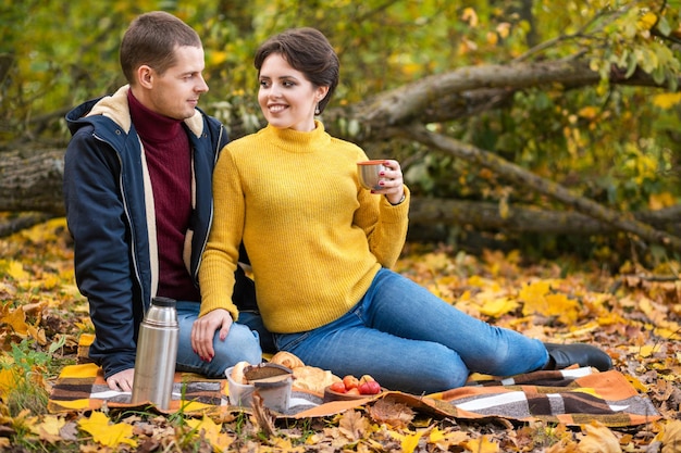 A beautiful young couple sits on a plaid in an autumn park drinks a hot drink from a thermos and hugs