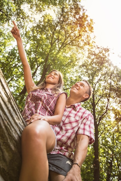 Красивая молодая пара сидит на упавшем дереве в лесу и наслаждается. Подруга показывает пальцем вдаль.