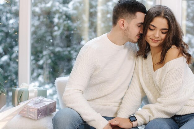 Beautiful young couple sitting in front of big window and fir tree its snow outside