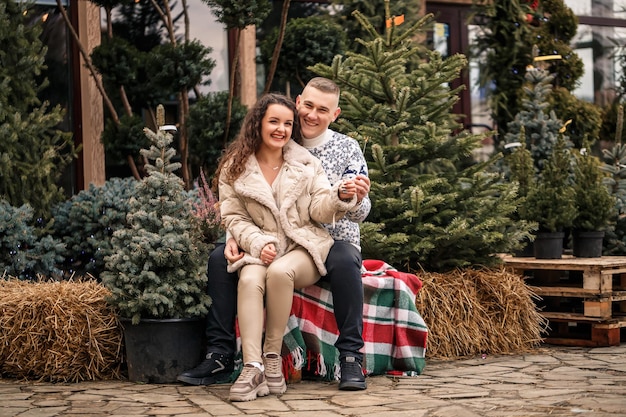 A beautiful young couple sitting near Christmas trees in white clothes walking near garlands Happy man and woman romance Christmas celebration fun love White sweater new year street outside