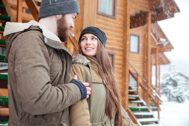 Beautiful young couple standing and looking at each other near wooden cottage in winter