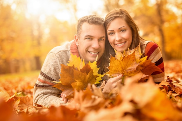 Beautiful young couple in sunny forest in autumn colors. They are lying on the fall meadow at embraced and holding yellow leaves. Looking at camera.