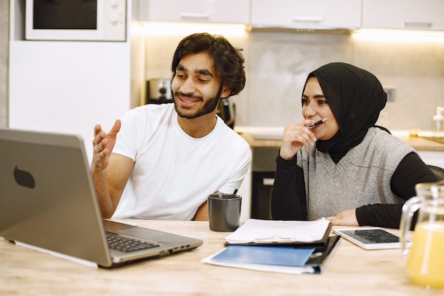 Beautiful young couple using a laptop, writing in a notebook, sitting in a kitchen at home. Arab girl wearing hidjab.