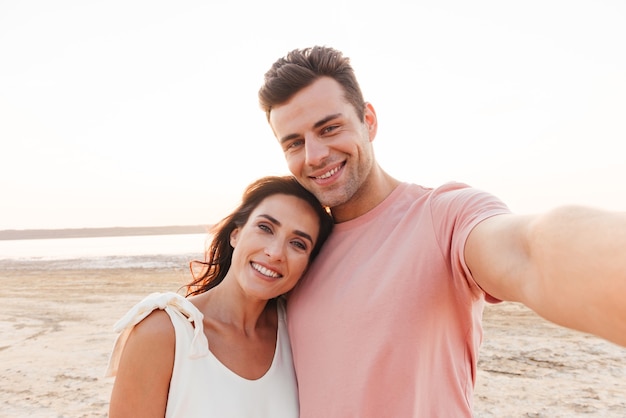 Beautiful young couple wearing summer clothing standing at the beach, taking a selfie, hugging