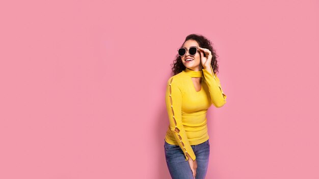 Beautiful young curly caucasian girl in a yellow outfit who is smiling and posing on pink background