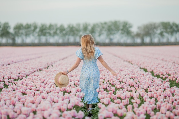 Beautiful young curly happy woman in pink tulip field in holland at sunset Stylish model with a light romantic dress in a blooming field