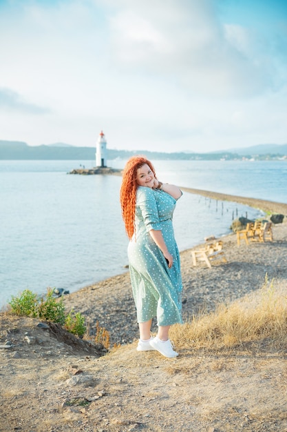 A beautiful young curvy woman with long red hair stands against the background of an old lighthouse