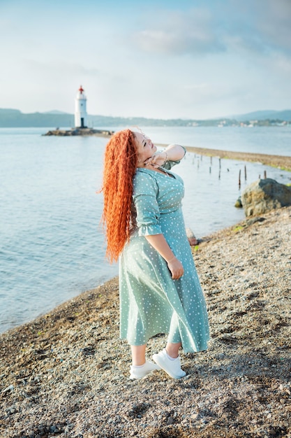 A beautiful young curvy woman with long red hair stands against the background of an old lighthouse