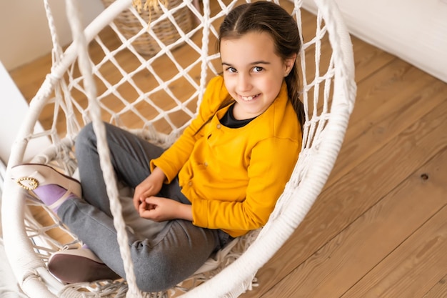 A beautiful young dark-haired girl relaxes in a swing. Cute brunette girl swinging in a woven chair at home. Looks at the camera.