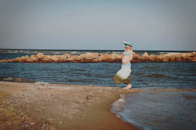 Beautiful young elegant pregnant blond woman in a blue hat walks on the beach, seashore at sunset.