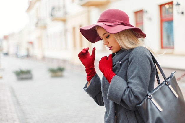 Bella giovane donna elegante in un cappello alla moda e un cappotto grigio con una borsetta sulla strada