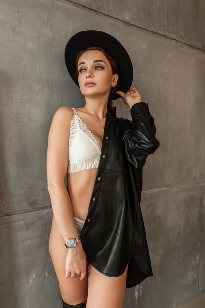 Beautiful young elegant woman with sexy body in white lingerie and black shirt with vintage hat stands near a gray wall indoors