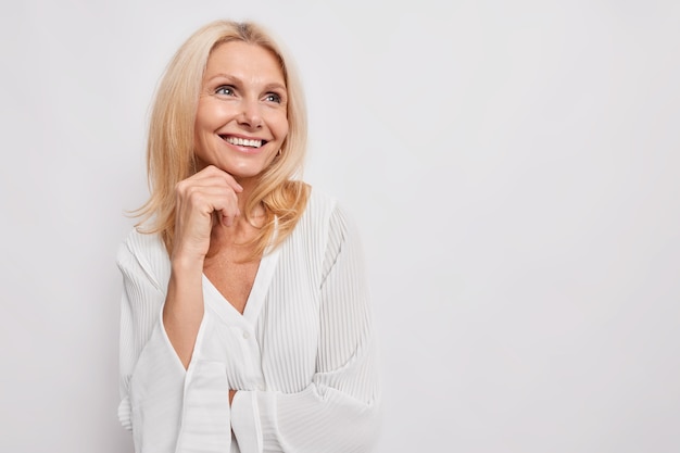 Photo beautiful young european woman of middle age smiles gently keeps hand under chin looks away with dreamy expression wears silk blouse isolated over white wall copy space for advertisement