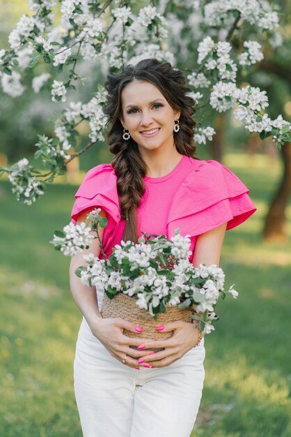 A beautiful young European woman with braids in the spring in the park holds a basket of flowers in a blooming orchard of apple trees