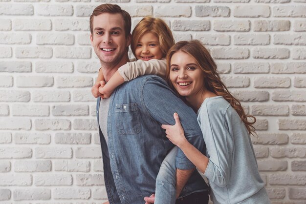 Beautiful young family hugging looking in camera and smiling while standing against white brick wall