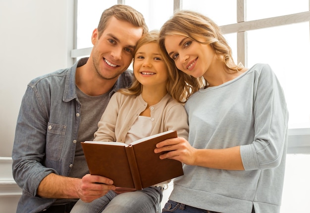 Photo beautiful young family looking in camera, smiling and holding a book while sitting near the window at home