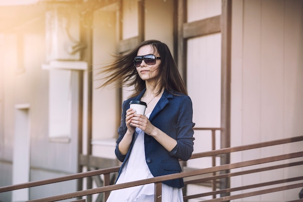 美しい若いファッションの女の子は、持ち帰り用のコーヒーを手にカフェから来ています