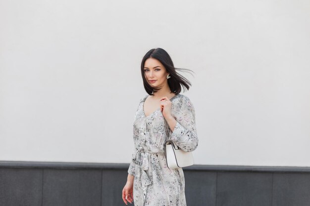 Beautiful young fashion girl in stylish elegance dress with flowers print with trendy fashion handbag stands near a white wall on summer windy day